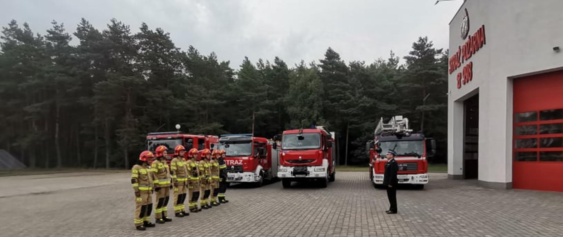 Na zdjęciu widać mężczyzn, strażaków w ubraniach specjalnych ustawionych w szeregu, przed nimi stoi strażak w ubraniu galowym. za nimi stoją wozy bojowe i podnośnik .