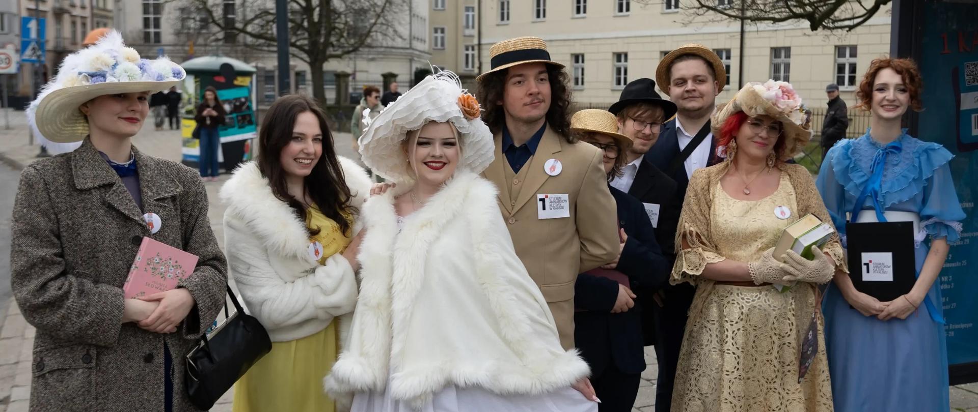 Grupa słuchaczy Studium Animatorów Kultury pozuje na przystanku w centrum Kalisza. Uczestnicy mają stylizowane stroje i kapelusze, a część z nich trzyma książki i materiały związane z poezją.