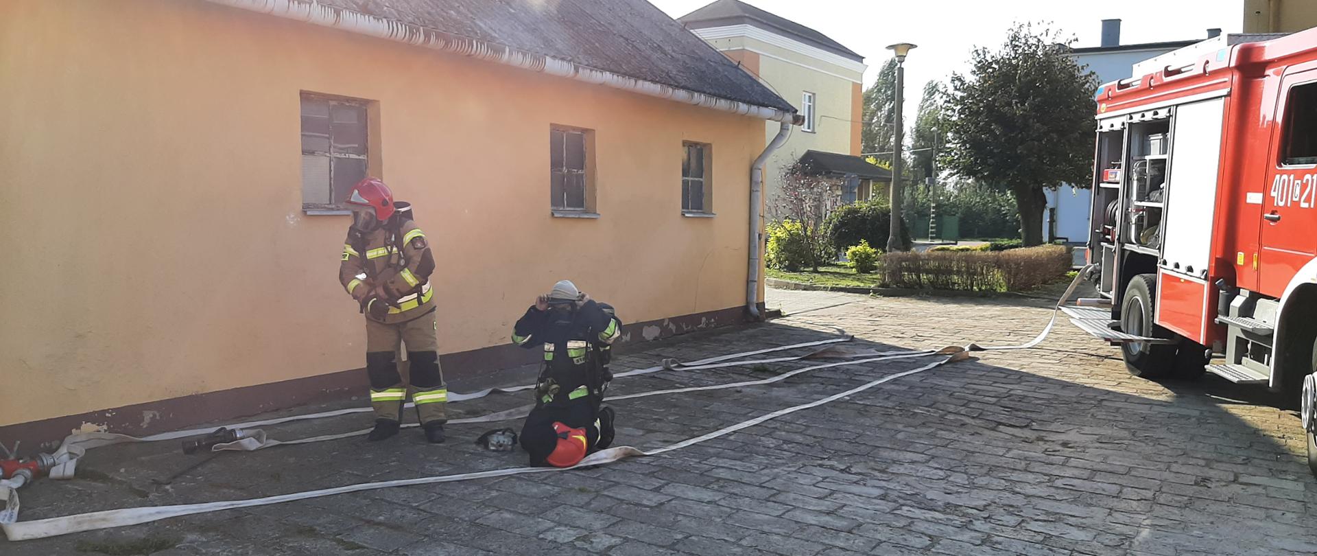 Zdjęcie przedstawia dwóch strażaków przygotowujących się na szarej kostce do akcji gaśniczej. Jeden ubrany jest w strój specjalny koloru żółtego, na głowie ma czerwony hełm, dugi ubrany jest na czarno, na głowie założoną ma jasną kominiarkę. Oboje posiadają na twarzach maski chroniące przed dymem. Za nimi po lewej stronie znajduje się pomarańczowy budynek z dachem pokrytym szarym eternitem. W centralnej części zdjęcia w oddali widać kolejny jasny budynek oraz szarą latarnię uliczną i zielone krzewy i drzewo. Po prawej stronie stoi czerwony samochód pożarniczy z otwartymi skrytkami na sprzęt strażacki. Od niego w kierunku strażaków ciągną się jasne węże pożarnicze.