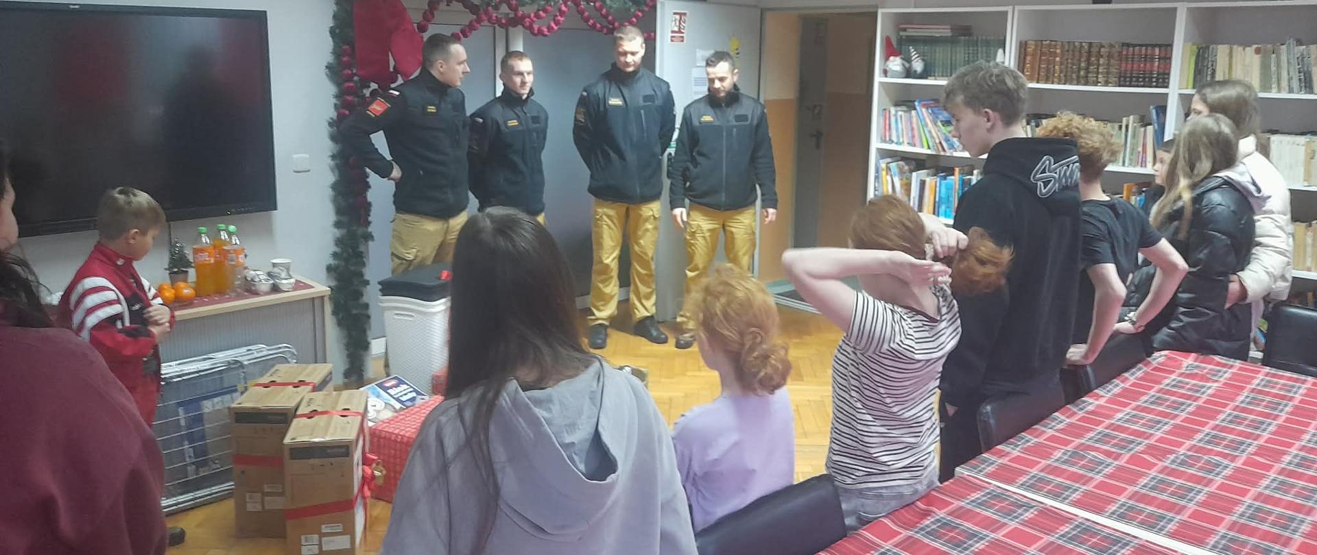 Strażacy w umundurowaniu służbowym stoją w świetlicy naprzeciwko grupy dzieci zgromadzonych przy stole. W pomieszczeniu znajdują się paczki z darami oraz świąteczne dekoracje