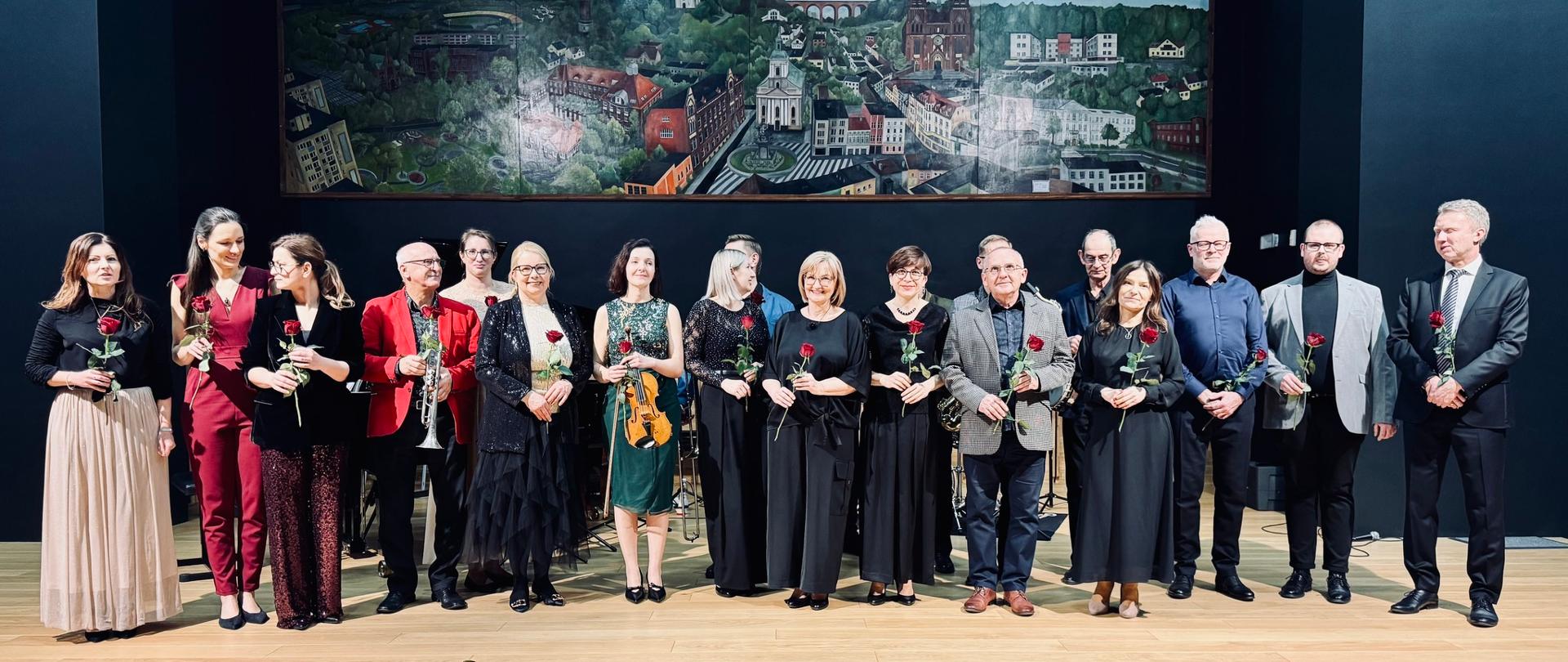 Na zdjęciu widać grupę kilkunastu nauczycieli stojących na scenie w jednym rzędzie. Wszyscy są elegancko ubrani i trzymają w dłoniach czerwone róże. W tle znajduje się duży obraz przedstawiający panoramę miasta z katedrą i zabudową miejską. Scena ma ciemne tło, a podłoga jest drewniana. Jest to pamiątkowe zdjęcie, wykonane po koncercie pedagogów.