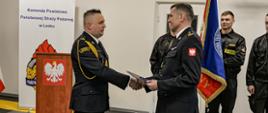 Zdjęcie przedstawia dwóch strażaków w mundurach wyjściowych - Komendanta Powiatowego Państwowej Straży Pożarnej w Lesku oraz Zastępcę Dowódcy Jednostki Ratowniczo – Gaśniczej w Lesku stojących na tle roll-upu Komendy Powiatowej Państwowej Straży Pożarnej w Lesku oraz mównicy. Komendant Powiatowy Państwowej Straży Pożarnej w Lesku ściska dłoń Zastępcy Dowódcy Jednostki Ratowniczo – Gaśniczej w Lesku.