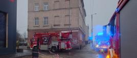 Zdjęcie w ciągu dnia. Zachmurzenie duże. Widać budynki oraz drogę publiczną. W pobliżu samochody ratowniczo-gaśnicze w kolorze czerwonym, posiadają sygnalizację ostrzegawczą.