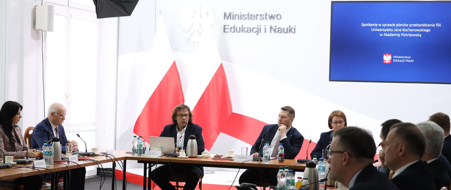 Grupa osób siedzących przy stole. Na stole napoje, termosy i mikrofony. Na ścianie ekran z napisem Spotkanie w sprawie planów przekształcenia filii Uniwersytetu Jana Kochanowskiego w Akademię Piotrkowską
