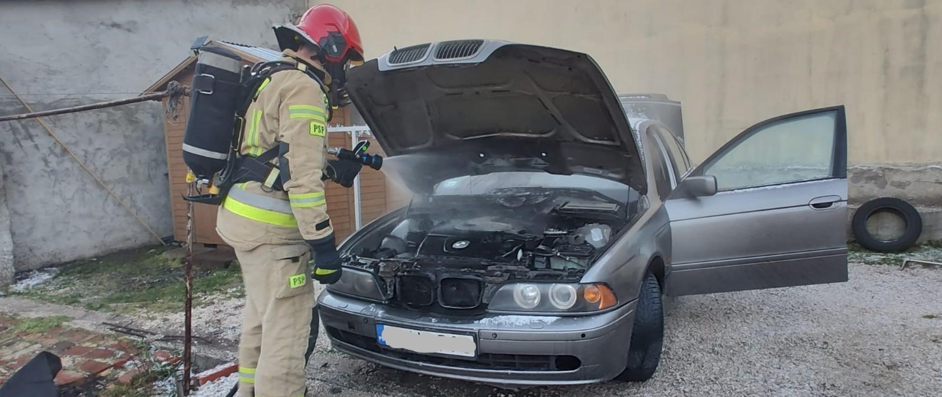 Zdjęcie przedstawia samochód osobowy z otwartą pokrywą silnika. Z komory silnika wydostaje się dym i para wodna. Przed autem stoi strażak podający prąd wody.