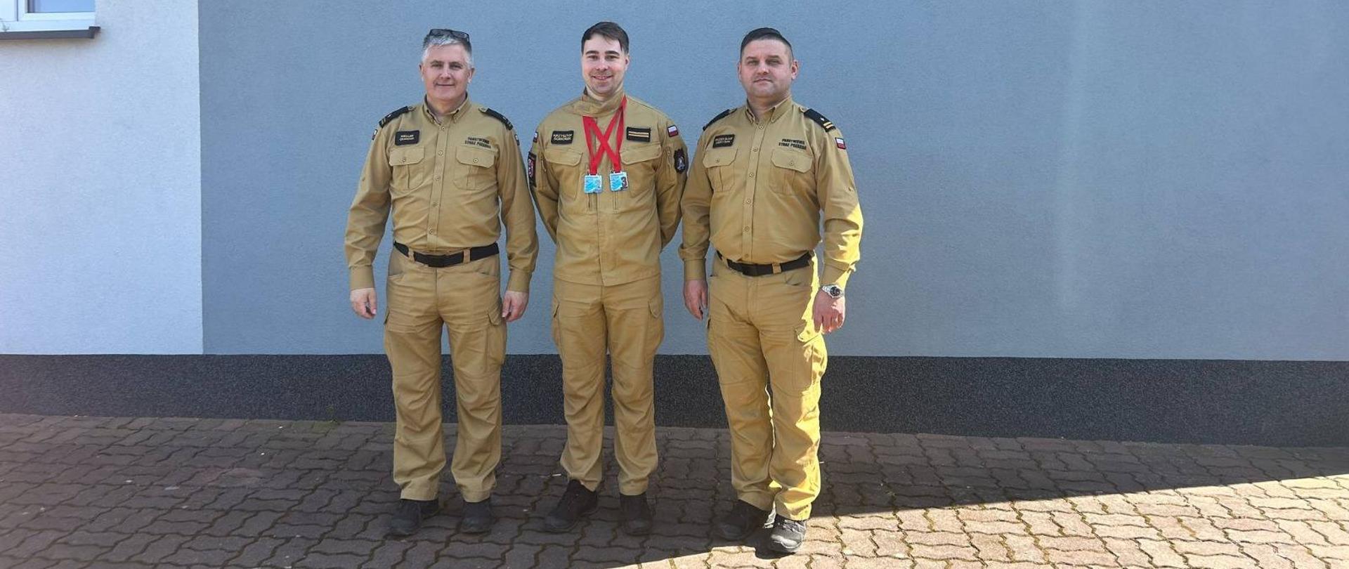 Na zdjęciu widać trzech strażaków, którzy pozują do zdjęcia. Wszyscy ubrani są w piaskowe mundury Państwowej Straży Pożarnej. Strażak w środku na szyi zawieszone ma dwa medale. W tle widać ścianę i czerwony napis Komenda Powiatowa Państwowej Straży Pożarnej w Łęczycy. Jest widno. 