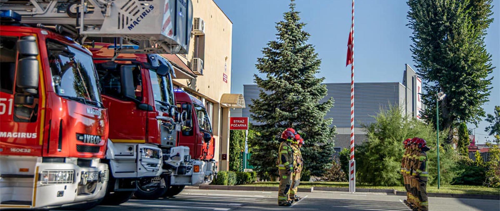 Strażacy PSP w umundurowaniu bojowym stojący przed samochodami oddają hołd strażakowi KG PSP, który zginął podczas służby.
