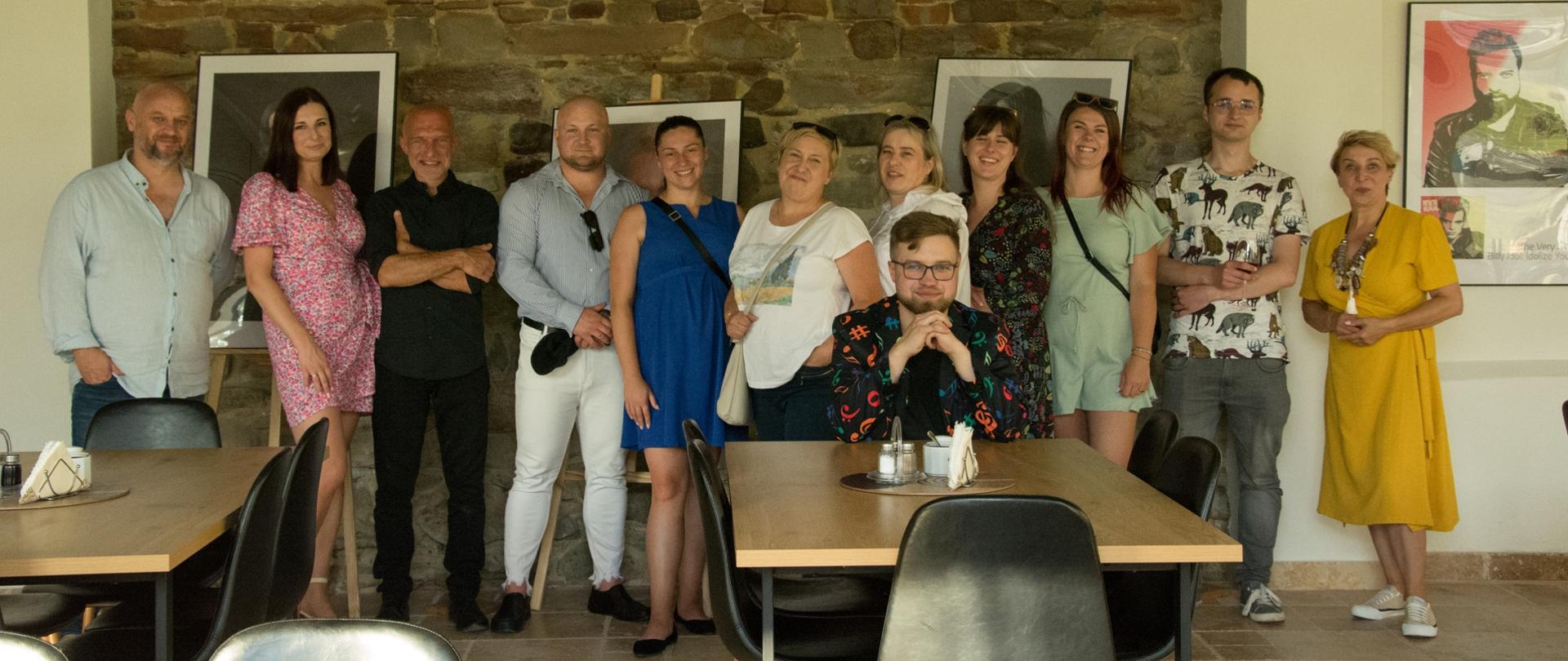 Grupa ludzi podczas wystawy fotograficznej, stoją na tle zdjęć i kamiennego muru.