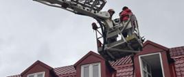 Zdjęcie wykonane w pochmurny dzień. Centralny punkt zdjęcia stanowi kosz podnośnika i znajdujące się w nim osoby. Strażak oraz osoby ewakuowane z płonącego budynku.