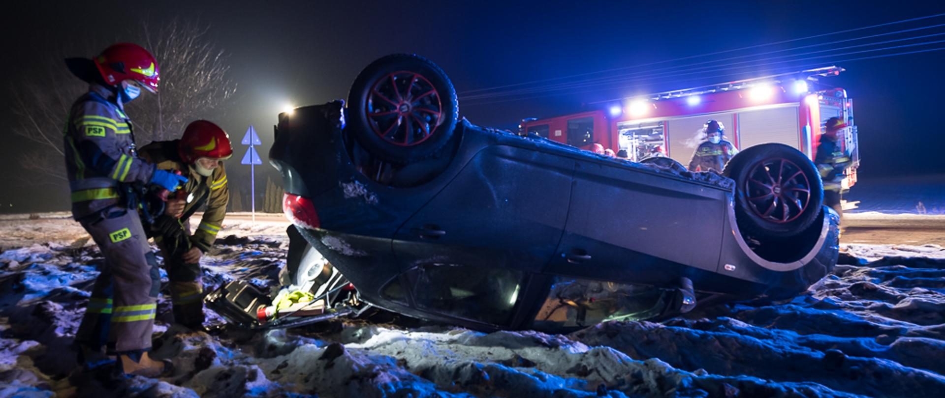 Samochód osobowy leżący na dachu na ośnieżonym polu. Do bagażnika zagląda dwóch strażaków.
