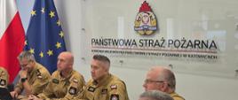 Na zdjęciu w sali konferencyjnej Komendy Wojewódzkiej Państwowej Straży Pożarnej w Katowicach siedzi przy stołach siedmiu funkcjonariuszy Państwowej Straży Pożarnej w piaskowych mundurach służbowych. Po środku zastępcy Śląskiego Komendanta Wojewódzkiego PSP – st. bryg. Roman Klecha i bryg. Zbigniew Dyk. Strażacy uczestniczą w spotkaniu lub wideokonferencji, skupieni przed dużym ekranem znajdującym się po lewej stronie kadru. Na stołach znajdują się laptopy, tablety, telefony, dokumenty i notatki. W tle widoczne są cztery flagi – dwie Polski i dwie Unii Europejskiej. Na ścianie umieszczono znak Państwowej Straży Pożarnej oraz tablicę z napisem: „PAŃSTWOWA STRAŻ POŻARNA. KOMENDA WOJEWÓDZKA PAŃSTWOWEJ STRAŻY POŻARNEJ W KATOWICACH”.