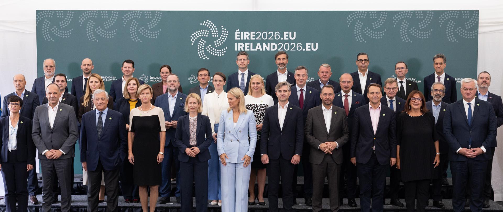 Grupowe zdjęcie uczestników spotkania Gymnich w Irlandii. Około 30 ministrów stoi w kilku rzędach przed ścianką z napisem „ÉIRE2026.EU IRELAND2026.EU”. Po bokach widoczne są flagi Unii Europejskiej i Irlandii.