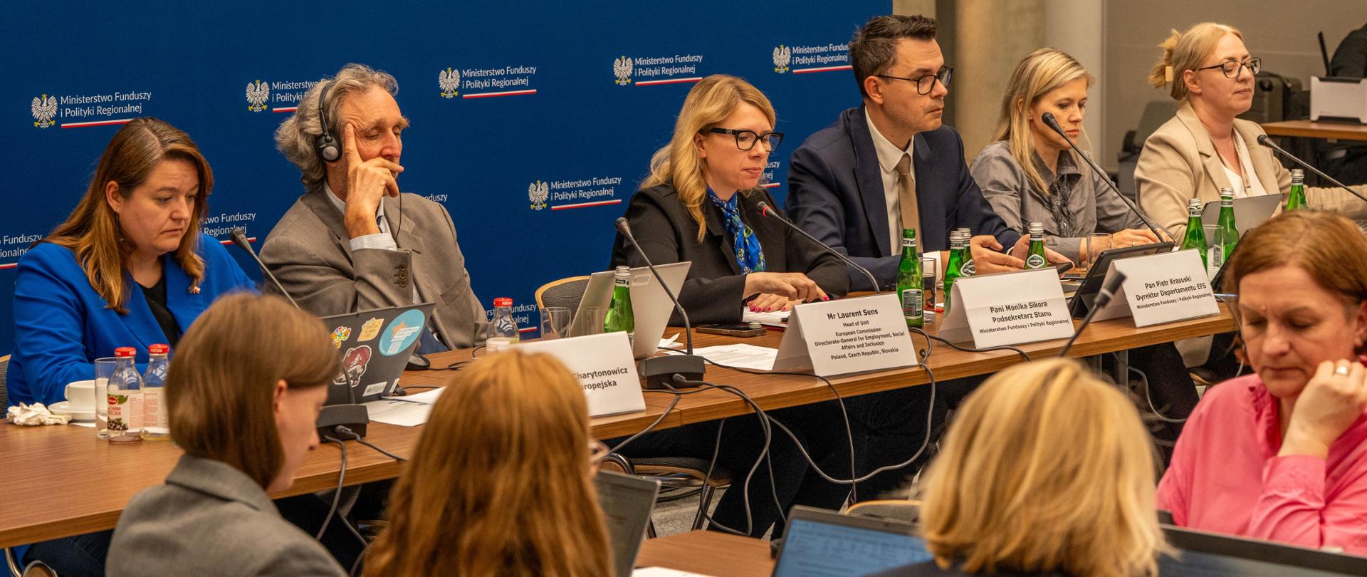 W sali konferencyjnej grupa osób siedzi przy stołach