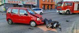 Na zdjęciu widzimy kolizję samochodu osobowego marki Opel Agila i skutera marki Yamaha. W wyniku kolizji w samochodu osobowego oraz skutera na drogę wyciekły płyny eksploatacyjne, które strażacy posypali sorbentem Compact. Samochód osobowy ma widoczne uszkodzenia przodu, wokół pojazdu znajdują się elementy karoserii. Bok skutera uległ deformacji, wokół niego znajdują się porozrzucane elementy karoserii.