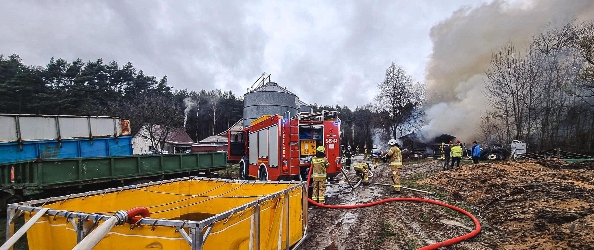 Strażacy gaszą pożar budynku gospodarczego. Na pierwszym planie widać rozstawiony zbiornik do wody.