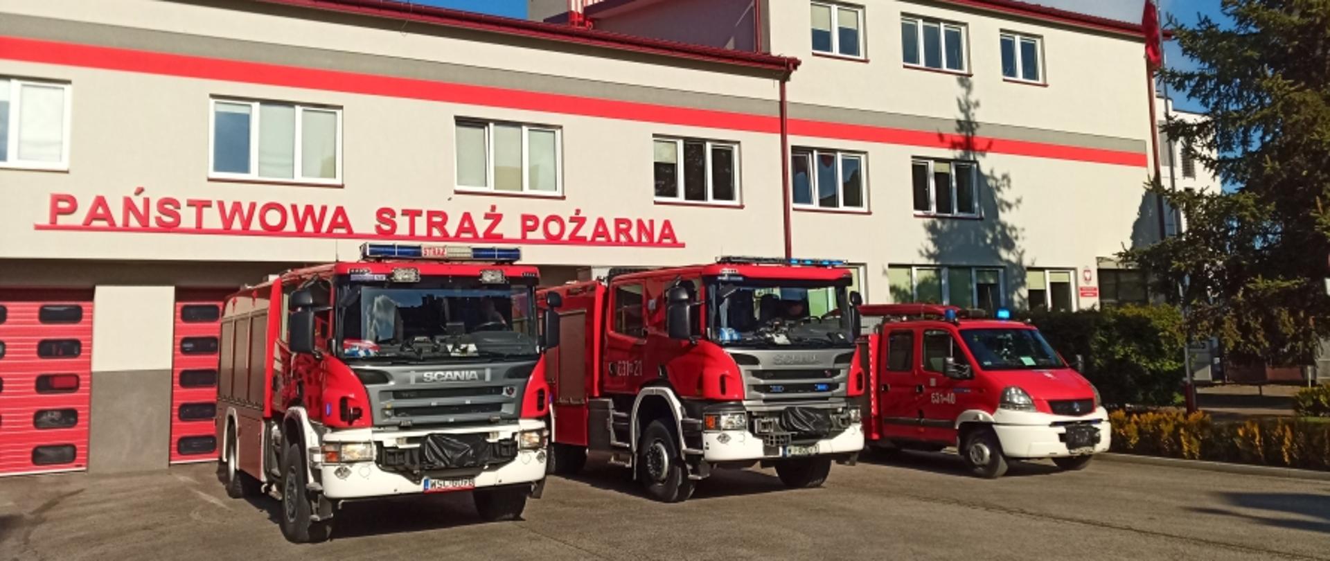 Na zdjęciu widoczne trzy samochody ratowniczo-gaśnicze przed garażami sochaczewskiej Jednostki Ratowniczo-Gaśniczej. Nad garażami widoczni napis Państwowa Straż Pożarna, a nad budynkiem wieża z napisem 112. Elewacja budynku w kolorach szaro-czerwonym, bramy garażowe czerwone.