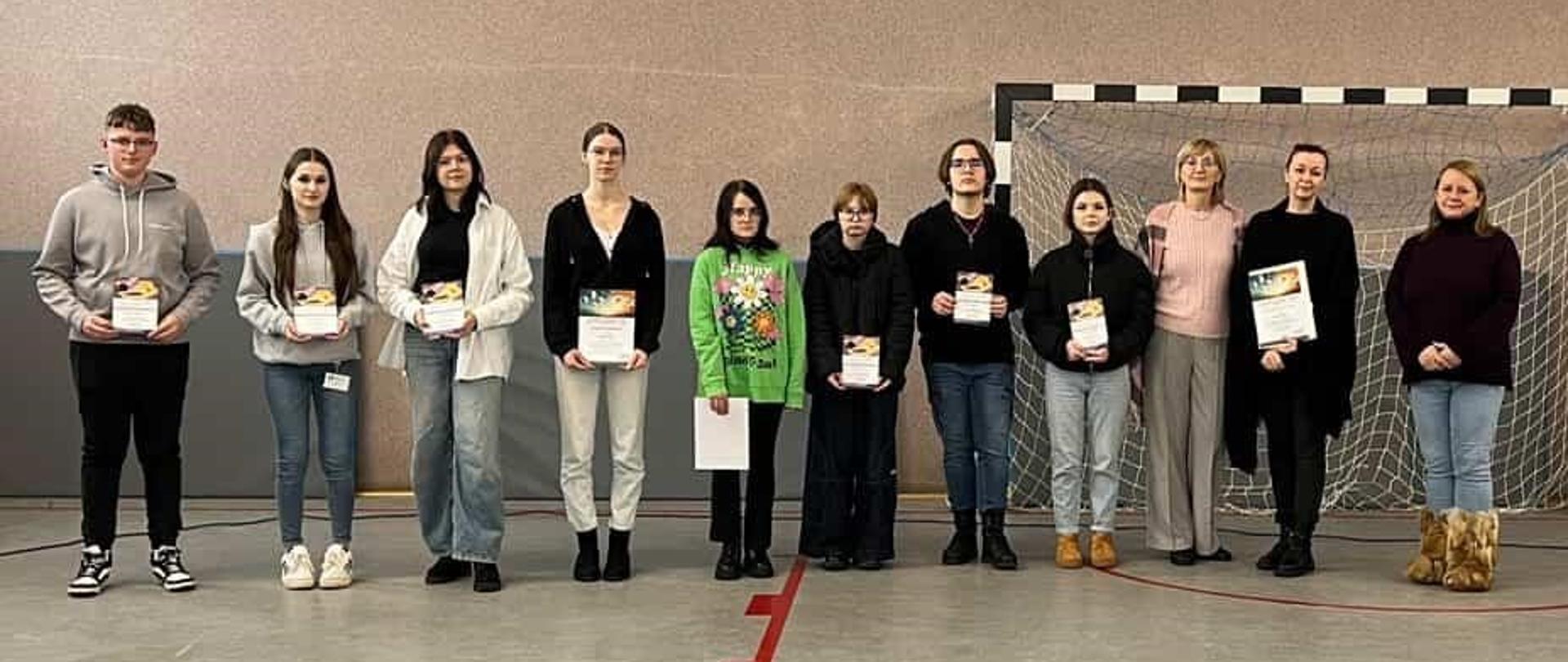 Grupa młodzieży i dorosłych stoi w rzędzie na hali sportowej, trzymając pamiątkowe dyplomy po zakończeniu szkolnego wydarzenia lub konkursu.