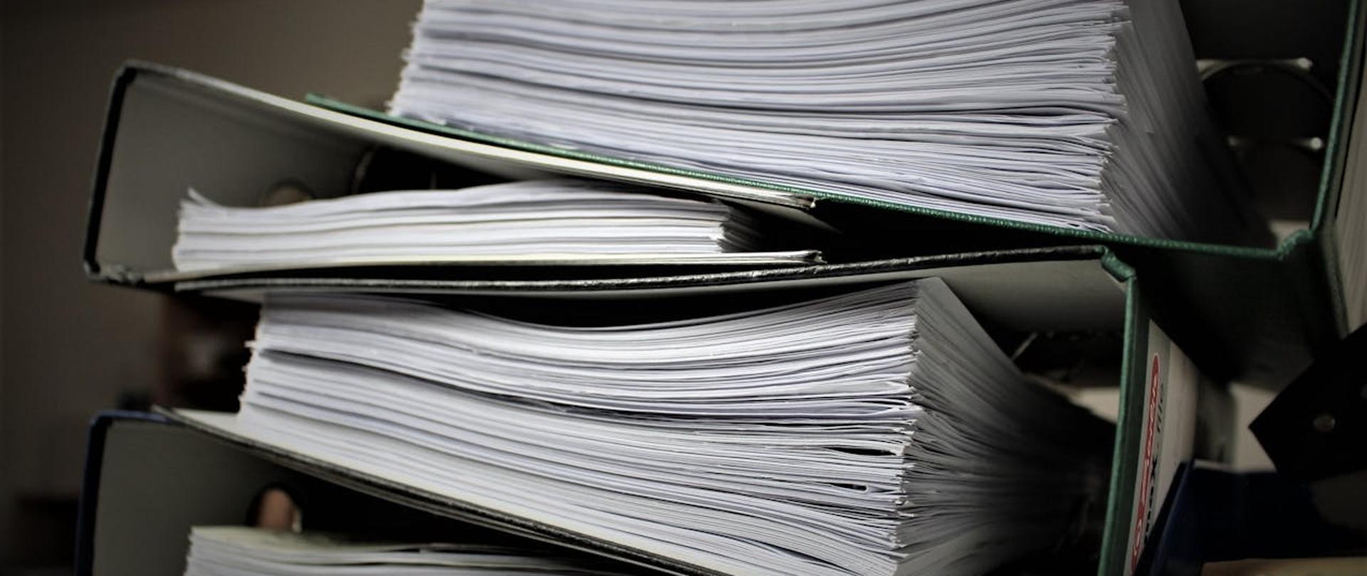 Pięć segregatorów z dokumentami. Segregatory są ułożone na sobie w taki sposób, że widać tylko górną krawędź i setki stron dokumentów.