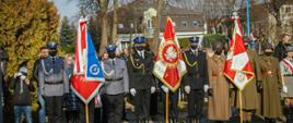 Poczty sztandarowe ustawione w szeregu, na pierwszym planie poczet sztandarowy komendy miejskiej policji złożony z policjantów w mundurach wyjściowych koloru szarego, poczet sztandarowy komendy miejskiej państwowej straży pożarnej w skierniewicach złożony z trzech strażaków w mundurach wyjściowych koloru czarnego. W tle pozostałe poczty sztandarowe złożone z żółnierzy i harcerzy. Fotografia wykonana w słoneczny dzień.