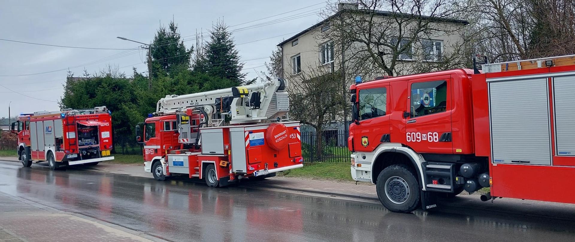 Zdjęcie przedstawia trzy czerwone wozy strażackie zaparkowane na mokrej nawierzchni drogi lub ulicy w pochmurny dzień. Na pierwszym planie po prawej stronie znajduje się duży wóz strażacki z numerem "609 68" widocznym na kabinie. W środku kadru stoi wóz, podnośnik hydrauliczny, z numerem "609 53" na boku. Za nim, w oddali po lewej stronie, widać trzeci, mniejszy wóz strażacki. Tło zdjęcia stanowią budynki mieszkalne z jasnymi elewacjami oraz drzewa i krzewy z widocznymi gałęziami, co sugeruje wczesną wiosnę lub jesień. Niebo jest szare i zachmurzone, a mokra nawierzchnia drogi odbija światło, co wskazuje na niedawny deszcz lub mżawkę. Całość obrazu ukazuje gotowość operacyjną jednostki straży pożarnej.