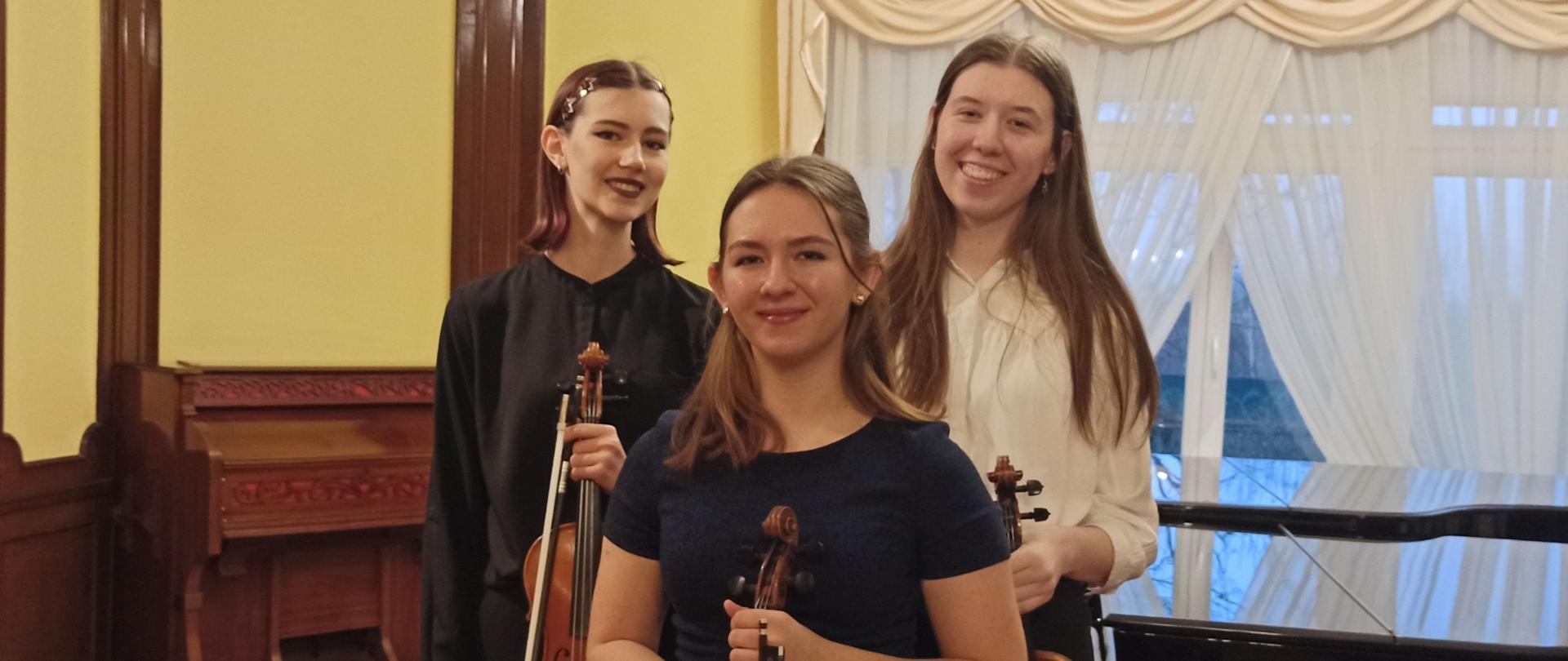 Zdjęcie kolorowe. Trzy uczennice ze skrzypcami pozują do zdjęcia. W tle fortepian, po lewej stronie zabytkowa fisharmonia.