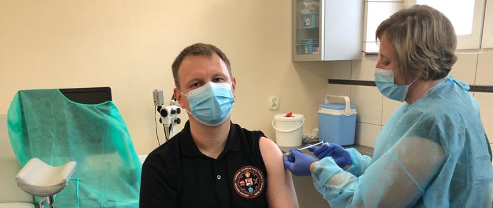 Na zdjęciu gabinet zabiegowy, strażak OSP siedzi na ławce ubrany w czarną koszulkę polo oraz spodnie dżinsowe. Po prawej stronie pielęgniarka ubrana w fartuch podaje szczepionkę w lewe ramię strażaka. Strażak oraz pielęgniarka mają założone na twarzy maseczki chirurgiczne.