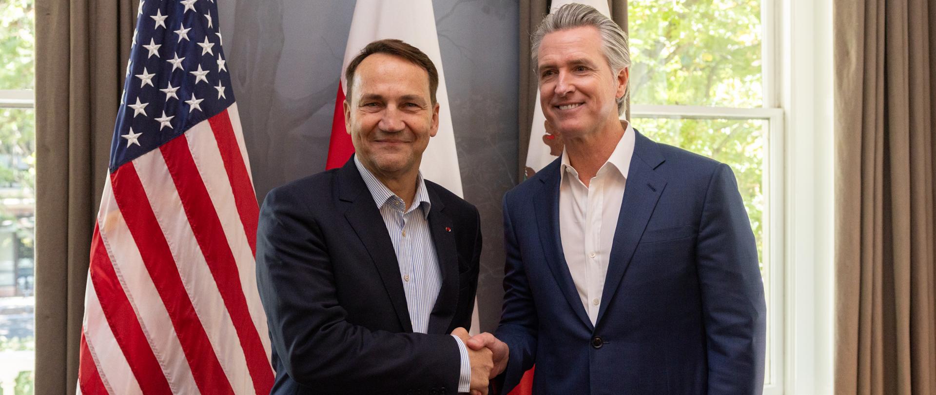 Na zdjęciu jest dwóch mężczyzn w garniturach. Za nimi jest okno i trzy flagi: USA, Polski i Kalifornii
