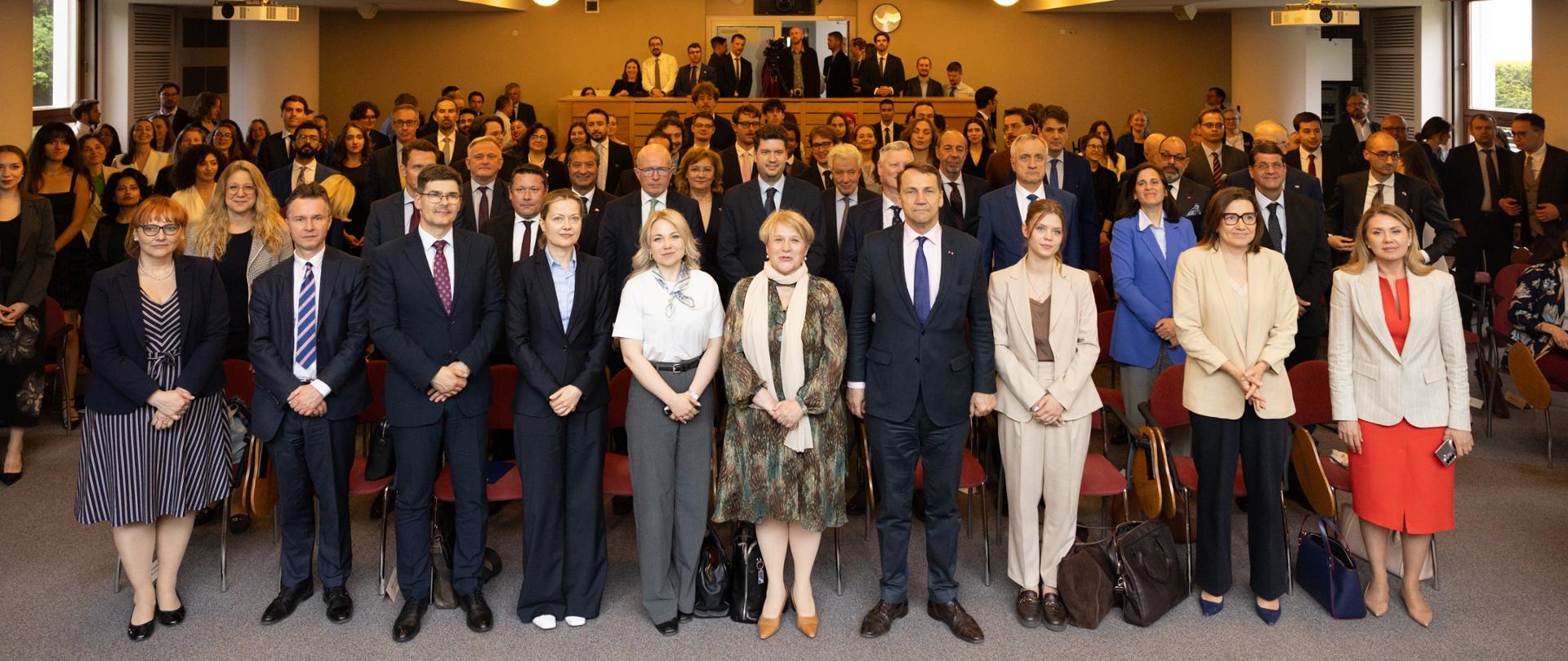 Na zdjęciu jest grupa kilkudziesięciu osób w eleganckich ubraniach. Patrzą w kierunku kamery. 