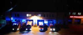Cztery samochody gaśnicze OSP Strzałkowo z włączonymi światłami błyskowymi przed budynkiem remizy z napisem, jednostka operacyjno techniczna widok z góry
