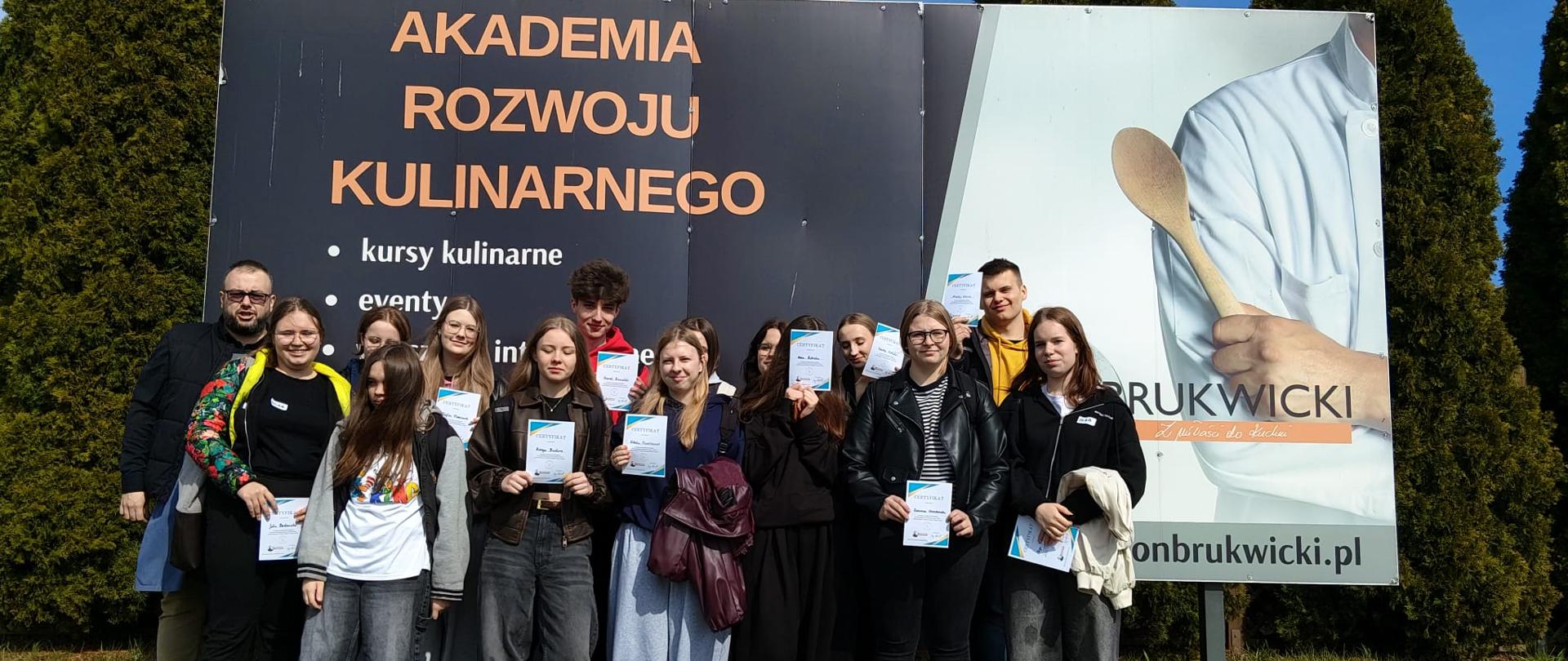 Grupa uczniów ZSCKR w Samostrzelu pozuje do pamiątkowego zdjęcia na tle billboardu Akademii Rozwoju Kulinarnego, dumnie prezentując otrzymane certyfikaty
