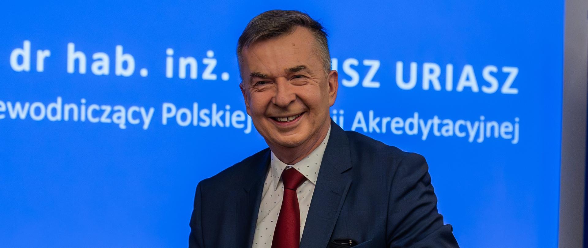 Minister Wieczorek stoi na tle wbudowanego w szarą ścianę wielkiego niebieskiego ekranu.