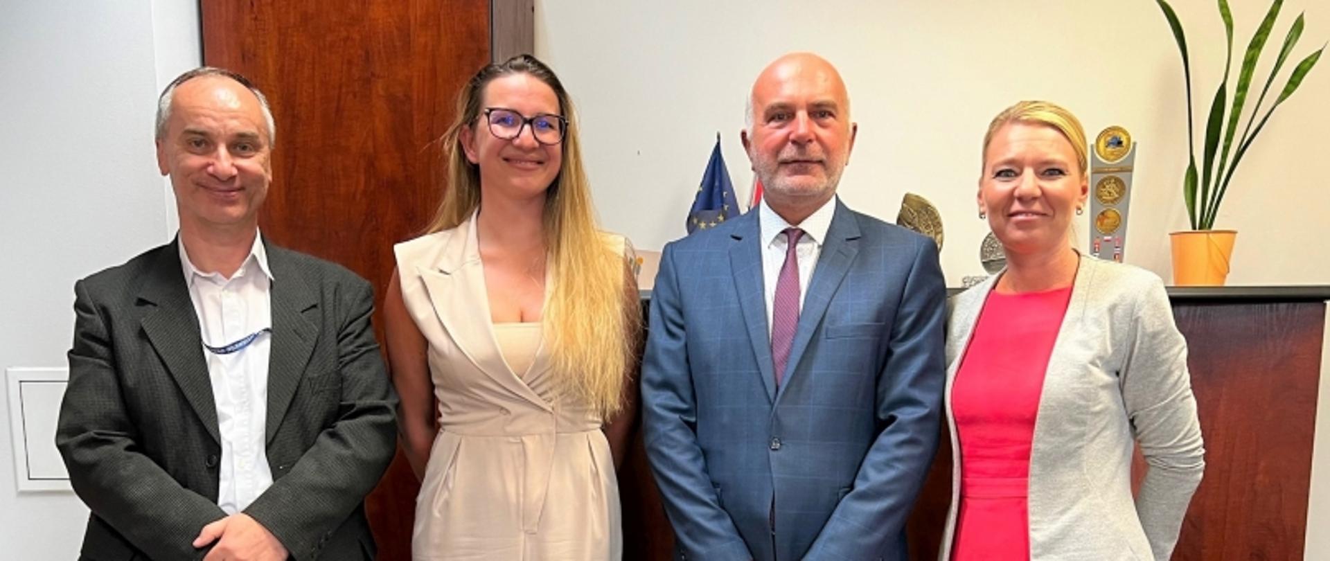 Cztery osoby stojące obok siebie w biurze; w tle flaga UE, drewniane drzwi i roślina doniczkowa.