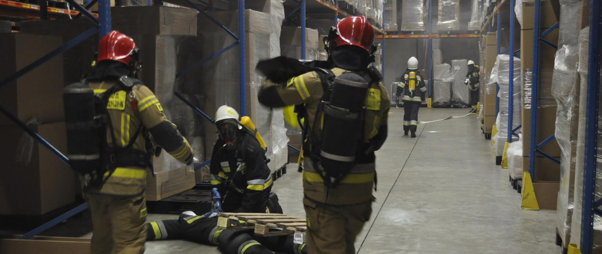 Zdjęcie przedstawia strażaków w hali magazynowej. Na pierwszym planie strażacy pozorują uwalnianie przygniecionego paletą manekina. W oddali dwaj strażacy rozwijają linie gaśnicze.