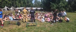 Strażak i policjantka prowadzą prelekcję dla licznej grupy uczestników obozu siedzących na trawie na otwartej polanie otoczonej lasem.