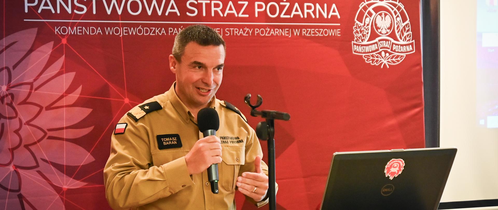 Mężczyzna w jasnobrązowym mundurze służbowym przemawia przy mównicy podczas podsumowania projektu „Bezpieczeństwo Ekologiczne”. W tle czerwona ścianka z logo Państwowej Straży Pożarnej w Rzeszowie. Przed nim laptop z naklejką przedstawiającą hełm strażacki.