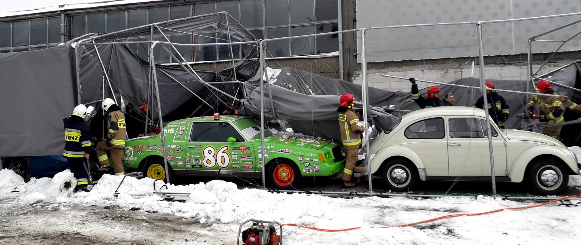 Zdjęcie przedstawia zniszczoną konstrukcję dużego brezentowego namiotu z usuniętym poszyciem. Wewnątrz namiotu stoją 2 samochody. Przy zabezpieczaniu uszkodzonej konstrukcji namiotu pracuje łącznie 6 strażaków, którzy używają narzędzi hydraulicznych