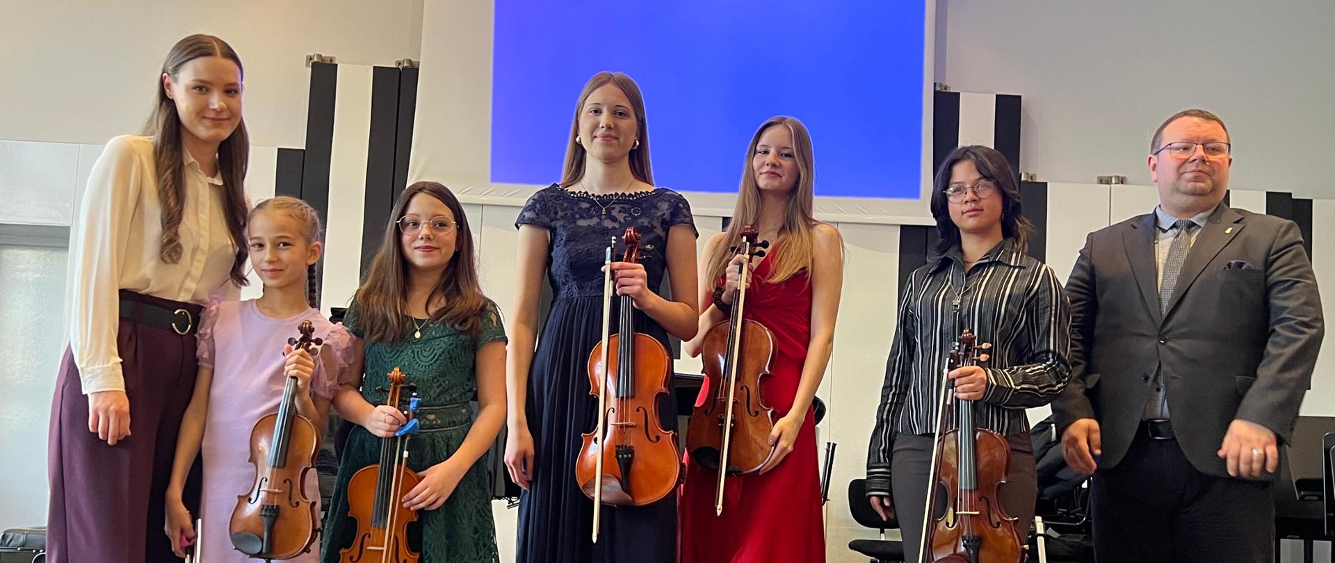 Pięć uczennic trzymających w rękach altówki stoi z kobietą i mężczyzną na scenie.