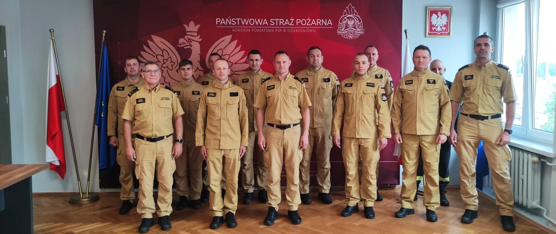 Fotografia grupowa przedstawiająca dziesięciu strażaków z Państwowej Straży Pożarnej. Mężczyźni stoją w dwóch rzędach w eleganckim pomieszczeniu biurowym, ubrani są w dopasowane, jasnobrązowe mundury służbowe z widocznymi odznakami i pagonami. Tło stanowi duża, bordowa ściana (backdrop), na której widnieje godło Polski (orzeł w koronie) oraz białe napisy: „PAŃSTWOWA STRAŻ POŻARNA” i poniżej, mniejszym drukiem: „KOMENDA POWIATOWA PSP W DZIERŻONIOWIE”. Po lewej stronie tła stoją na stojakach dwie flagi: flaga Polski (biało-czerwona) oraz flaga Unii Europejskiej (niebieska z gwiazdami). W pomieszczeniu widać parkiet ułożony w jodełkę, podwieszany sufit kasetonowy, okno po prawej stronie oraz ramię stołu konferencyjnego po lewej. Wszyscy strażacy patrzą prosto w obiektyw i mają neutralne lub lekko uśmiechnięte miny. Oświetlenie jest naturalne i równomierne.