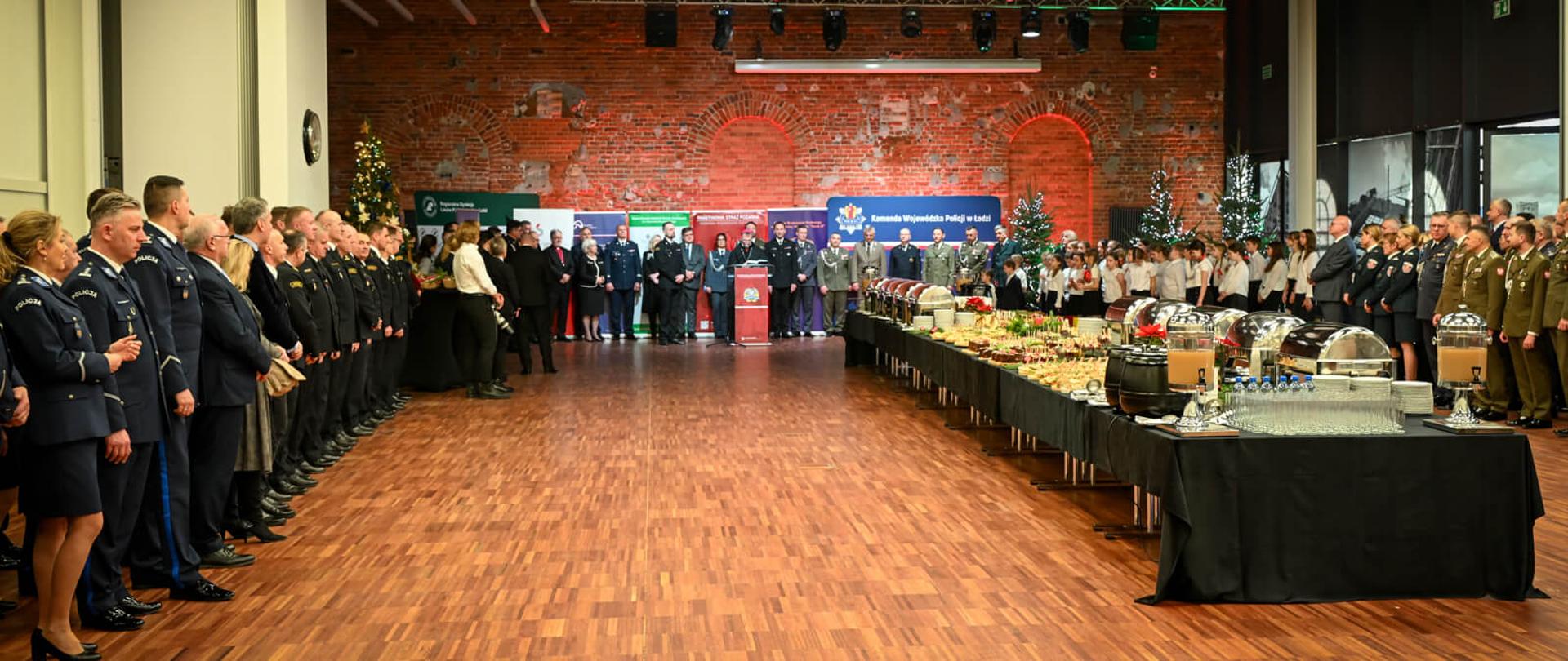 Szerokie ujęcie dużej, industrialnej sali z ceglanymi ścianami. Po lewej i prawej stronie stoją rzędy mundurowych, a na środku długie stoły z cateringiem. W głębi odbywa się oficjalna ceremonia.