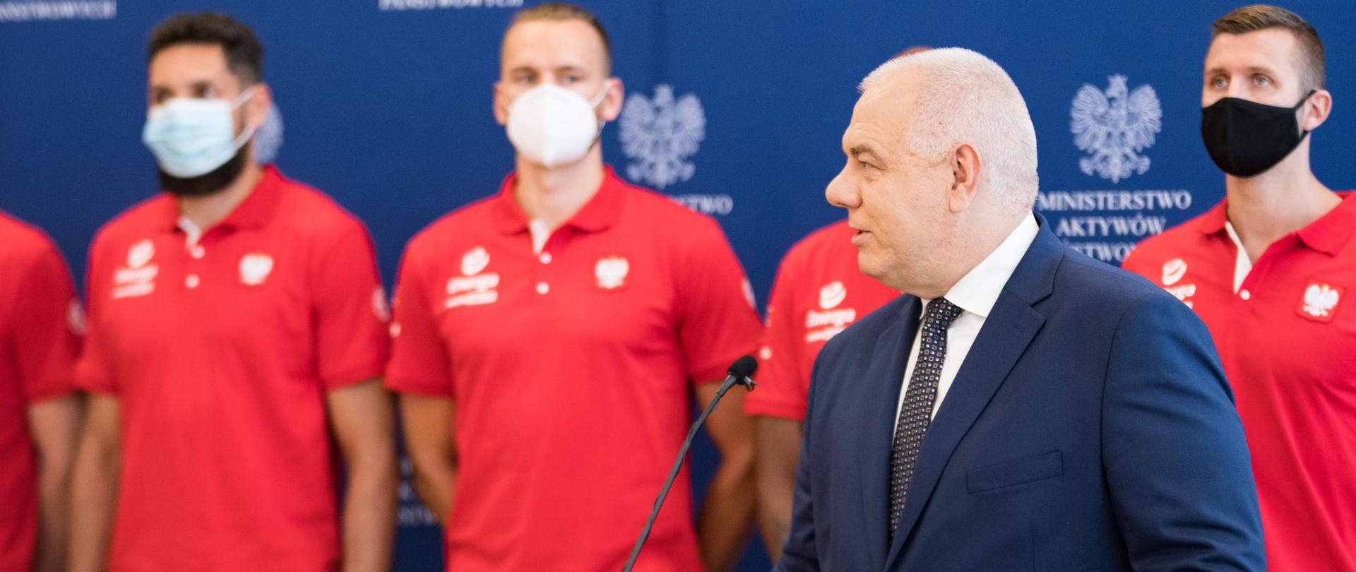 Wicepremier Jacek Sasin przemawia z mównicy. W tle koszykarze polskiej reprezentacji.