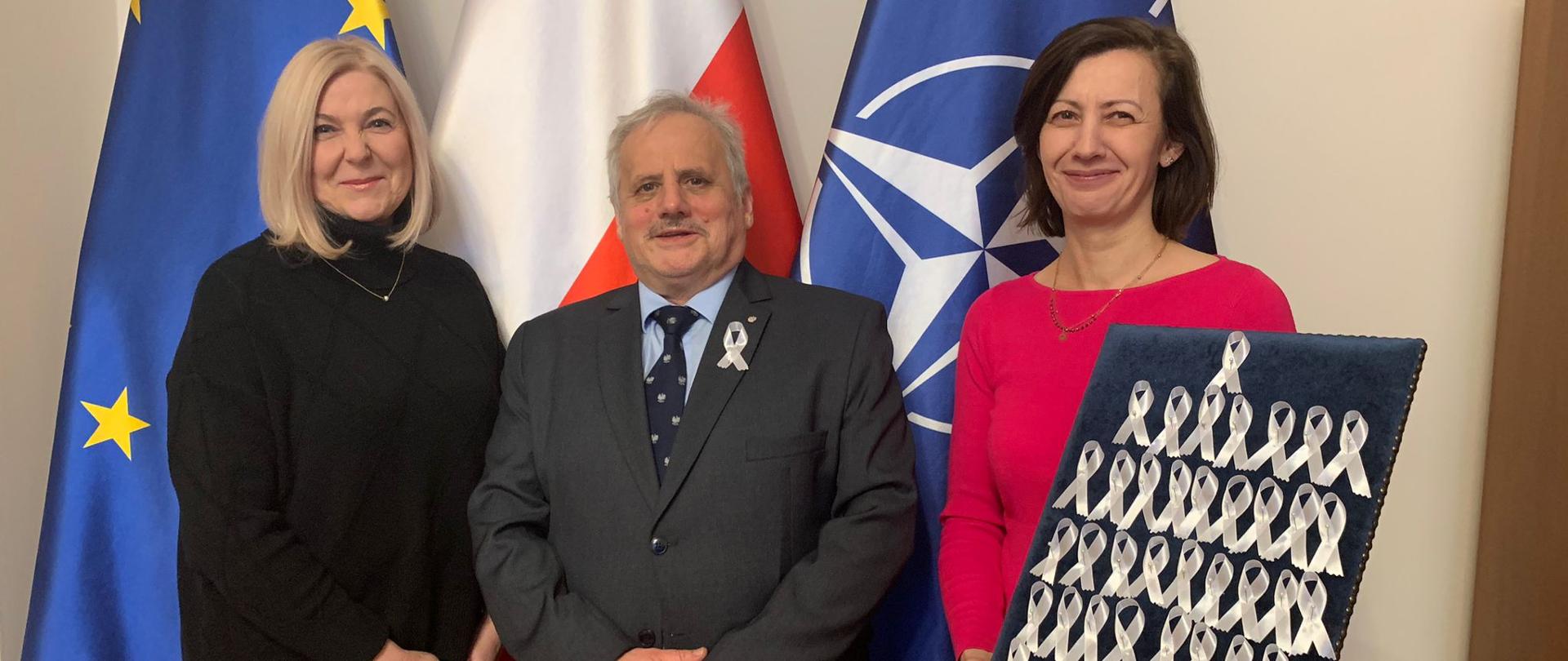 Dwie kobiety i jeden mężczyzna stoją ba tle flag, jedna kobieta trzyma tacę z białymi wstążkami. 