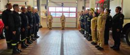 Garaż w remizie. Po lewej i prawej stronie stoją strażacy w dwuszeregu. W środku komendant i dowódca jednostki. W tle bramy garażowe remizy.