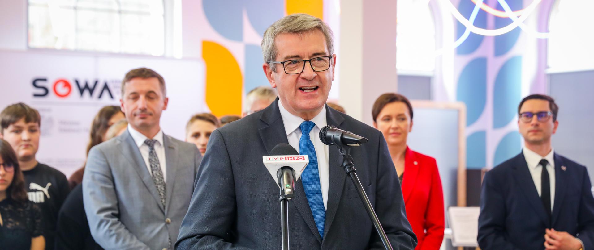 Wiceminister Wojciech Murdzek przemawia podczas otwarcie rybnickiej SOWY. Za nim stoją inni uczestnicy uroczystości.