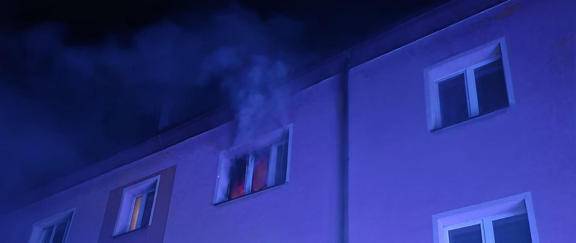 Zdjęcie przedstawia elewację wielopiętrowego budynku mieszkalnego nocą, oświetloną intensywnym, niebieskim światłem przypominającym błysk kogutów służb ratunkowych. Z okna na ostatnim piętrze wydobywa się gęsty dym, a wewnątrz pomieszczenia widać pomarańczową łunę ognia wskazującą na pożar mieszkania. Pozostałe okna bloku są ciemne lub słabo oświetlone, a cała scena uchwycona jest z niskiej perspektywy skierowanej ku górze.