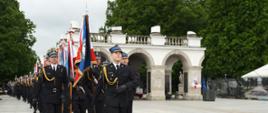 Kolumna funkcjonariuszy PSP w trakcie uroczystego przemarszu z Grobu Nieznanego Żołnierza. Na czele znajduje się poczet sztandarowy z widocznym sztandarem szkoły pożarniczej. Tło stanowi klasyczna architektura Grobu oraz zieleń Ogrodu Saskiego.