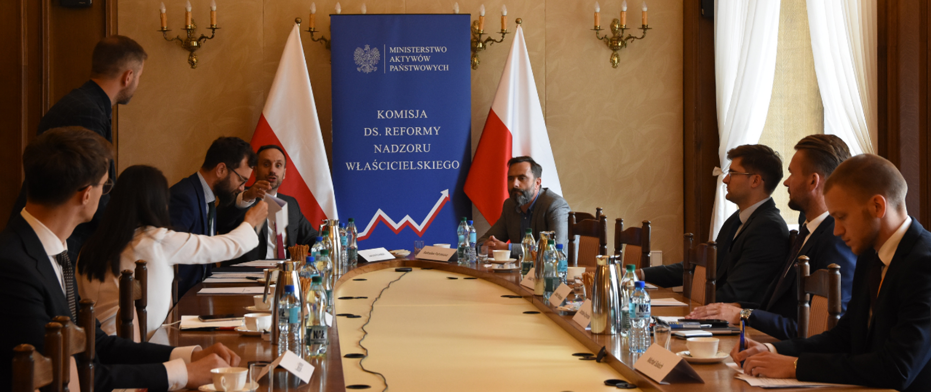 Uczestnicy spotkania siedzą przy owalnym stole. Wśród nich minister Janusz Kowalski. W tle ścianka z logotypami MAP i Komisji ds. Nadzoru Właścicielskiego oraz dwie flagi Polski
