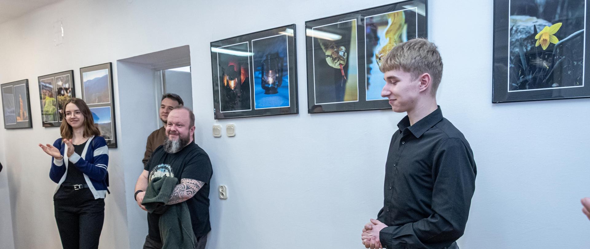 Zdjęcie przedstawia trzy osoby z publiczności zgromadzonej podczas wernisażu wystawy Perspektywa odbita w lusterku. Po prawej stronie stoi autor wystawy. W tle wiszą zdjęcia, które są zaprezentowane na wystawie. 