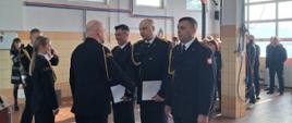 Na zdjęciu widać uroczystość w jasnym wnętrzu przypominającym halę lub garaż strażacki. Mężczyzna w galowym mundurze ściska dłoń jednemu z trzech funkcjonariuszy stojących w szeregu; pozostali trzymają w rękach dokumenty. Obok stoi kobieta również w mundurze. W tle znajdują się inni strażacy i osoby cywilne obserwujące wydarzenie. Pomieszczenie ma jasne ściany z kafelkami, duże okna, instalacje wentylacyjne pod sufitem oraz bramę garażową.
