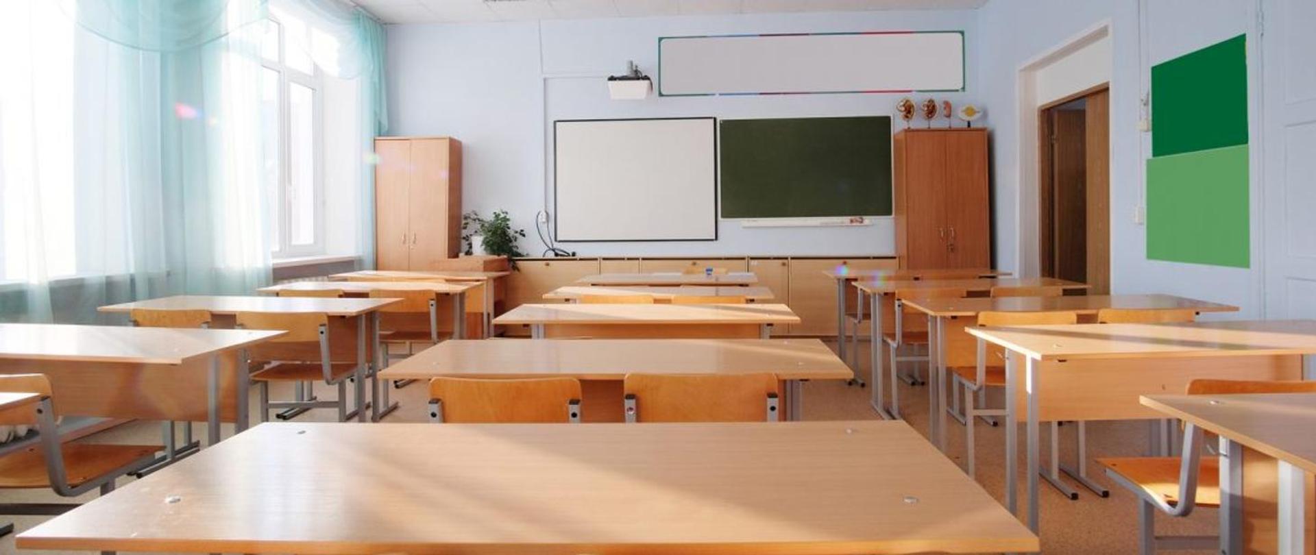 Zdjęcie przedstawia klasę szkolną, w której stoją ławki oraz krzesła, a także wiszą dwie tablice.