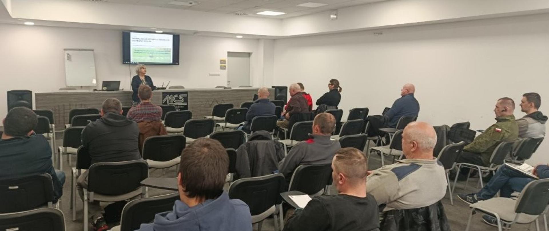Sala szkoleniowa: kilkanaście osób siedzi na krzesłach ustawionych w rzędach i patrzy w stronę przodu sali. Prelegentka stoi przy stole z laptopem, obok znajduje się ekran z wyświetloną prezentacją (tekst na slajdzie jest nieczytelny). Pomieszczenie jest dobrze oświetlone, z jasnymi ścianami i sufitem z panelami świetlnymi.